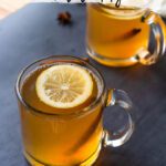 two clear mugs filled with an amber liquid and topped with a lemon slice and cinnamon stick; text overlay at the top of the image reads, 'non-alcoholic hot toddy'