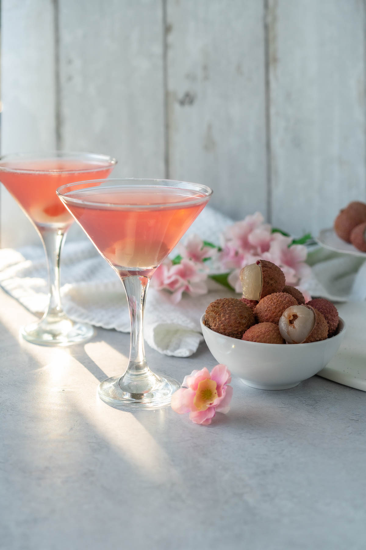 two martini glasses against a white background decorated with pale pink flowers and a bowl of lychee fruit; each glass is filled with a pale pink drink and garnished with a lychee