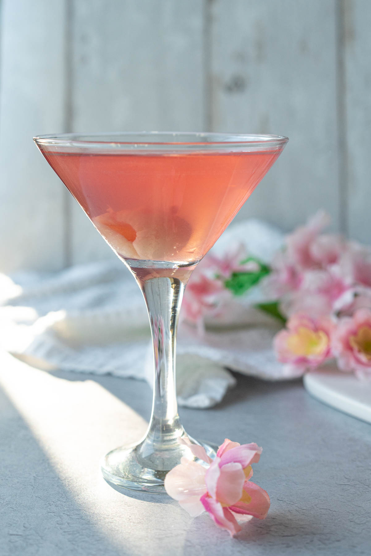 martini glass against a white background decorated with pale pink flowers; glass is filled with a pale pink drink and garnished with a lychee