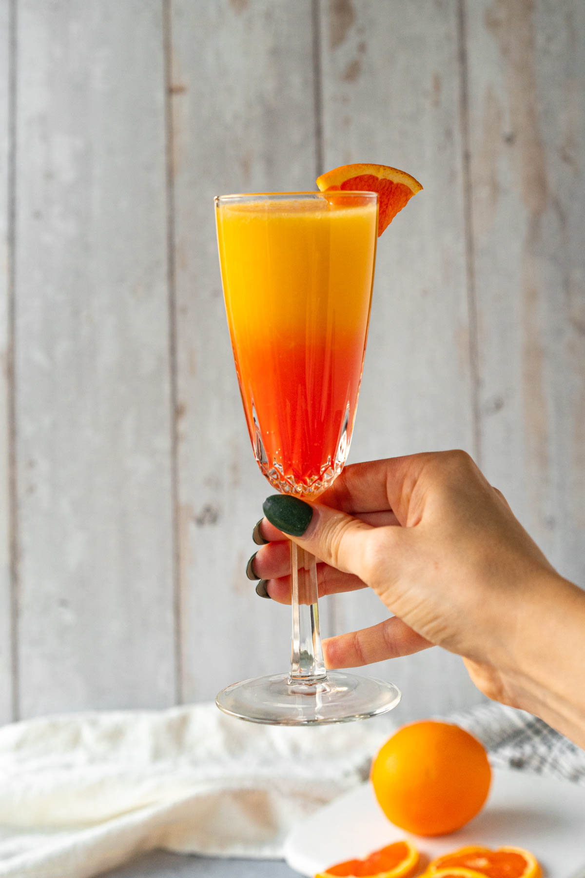 Hand holding a crystal champagne glass garnished with an orange slice. The glass is filled with orange juice and grenadine in a gradient fading from red-orange up to yellow-orange. 