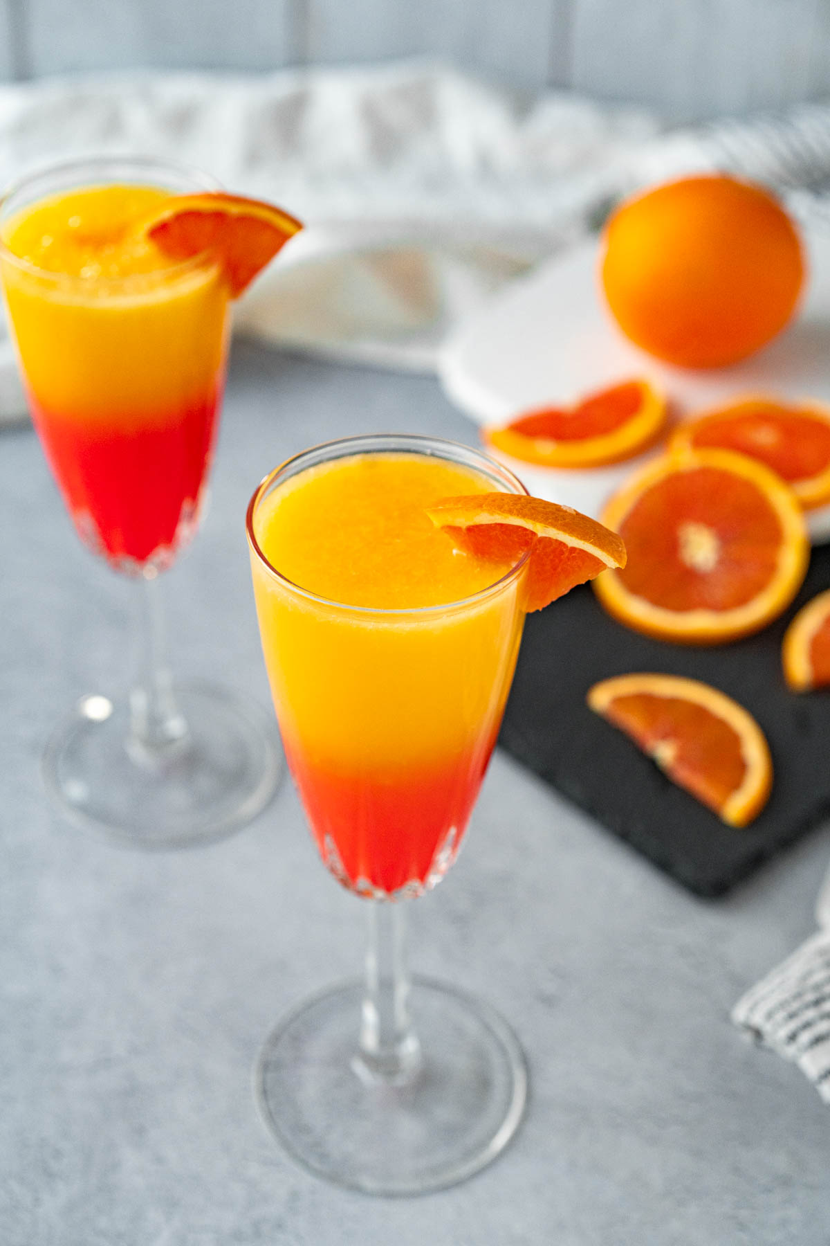 Closeup shot of two crystal champagne glasses, each garnished with an orange slice. The glasses are filled with orange juice and grenadine in a gradient fading from red-orange up to yellow-orange.