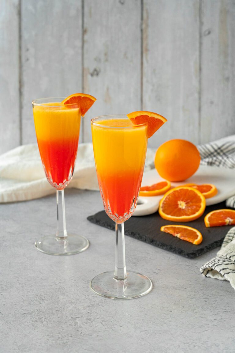 Two crystal champagne glasses, each garnished with an orange slice. The glasses are filled with orange juice and grenadine in a gradient fading from red-orange up to yellow-orange. A whole orange and orange slices are faded in the background.