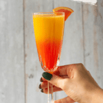 Hand holding a crystal champagne glass garnished with an orange slice. The glass is filled with orange juice and grenadine in a gradient fading from red-orange up to yellow-orange.
