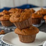 Two muffins stacked on a small white plate, with more muffins blurred in the background.