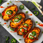 Top-down photograph of a white tray with four halved potatoes stuffed with melted cheese and topped with dollops of sour cream, sliced jalapeños, and sliced green onions. The tray is against a distressed gray background scattered with green onions and jalapeños.