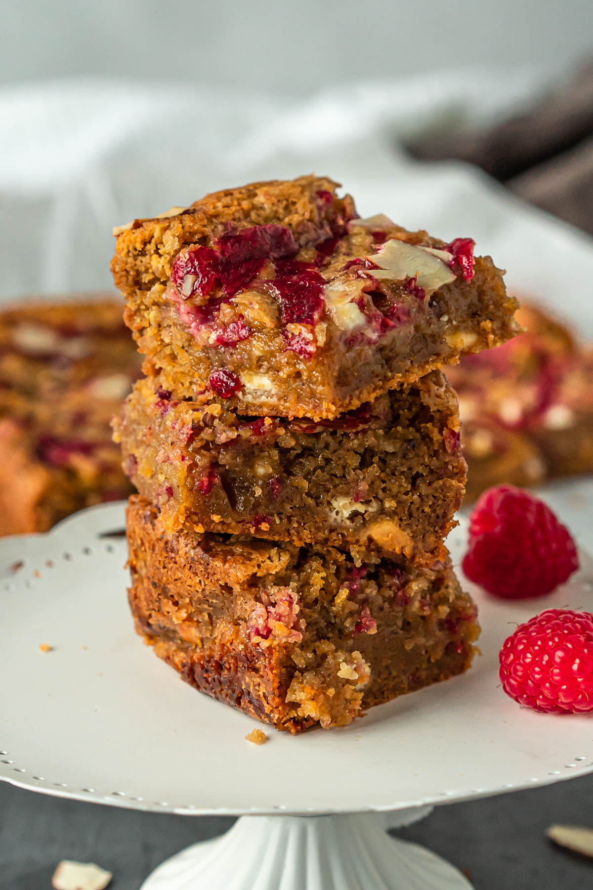 A stack of three square, golden-brown dessert bars drizzled with raspberry jam and studded with white chocolate and slivered almonds.