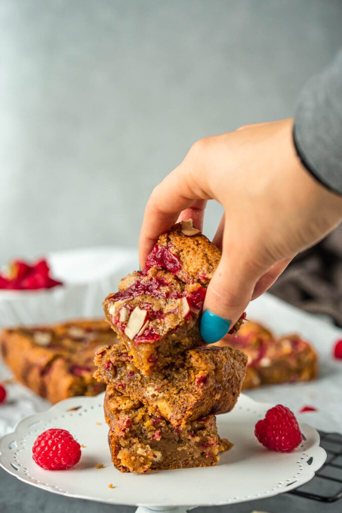 A stack of three square, golden-brown dessert bars drizzled with raspberry jam and studded with white chocolate and slivered almonds. A hand is grabbing the bar on top of the stack.